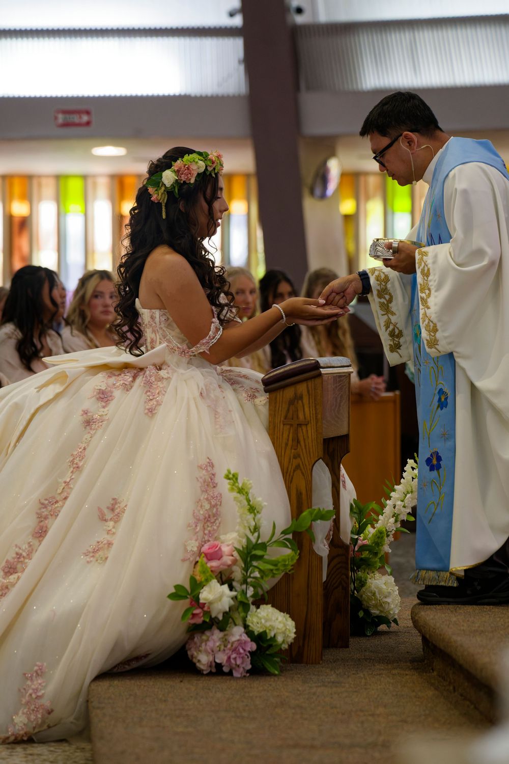 Priest offers communion to quinceañera girl in embroidered white ball gown with floral crown during church ceremony.