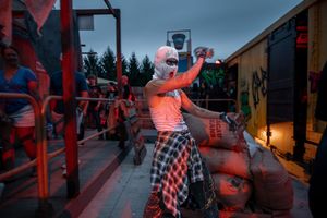 A masked performer in dramatic red lighting poses at an outdoor event with sandbags and graffiti in the background.