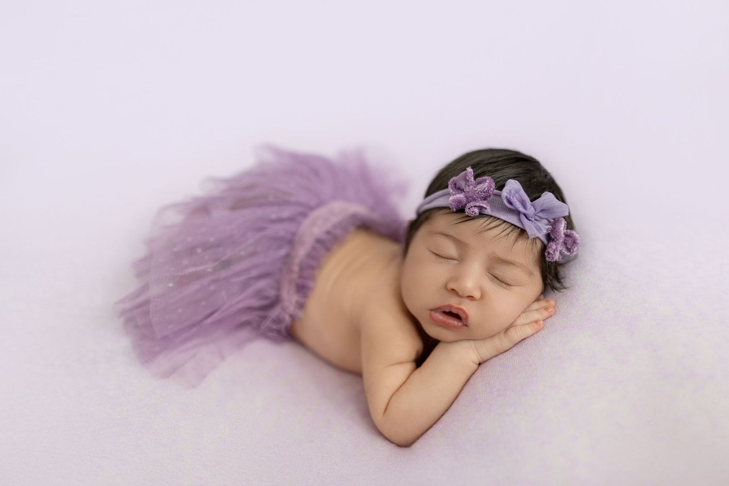 Sleeping newborn baby girl wearing a purple tutu skirt and floral headband on a white fluffy blanket.