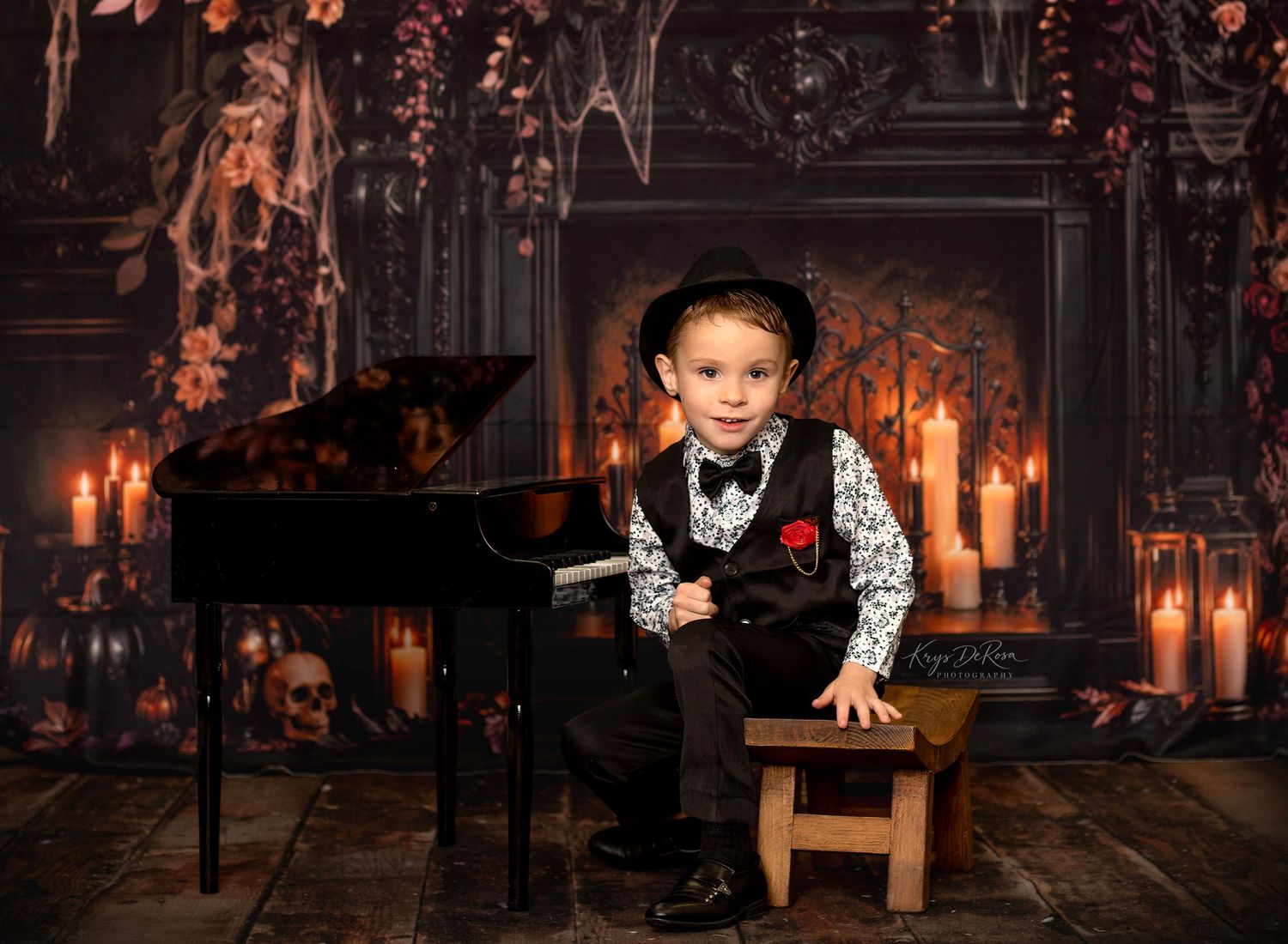 Young boy in a black hat, vest and bow tie sits on a wooden stool beside a grand piano in a spooky candlelit Halloween setting.