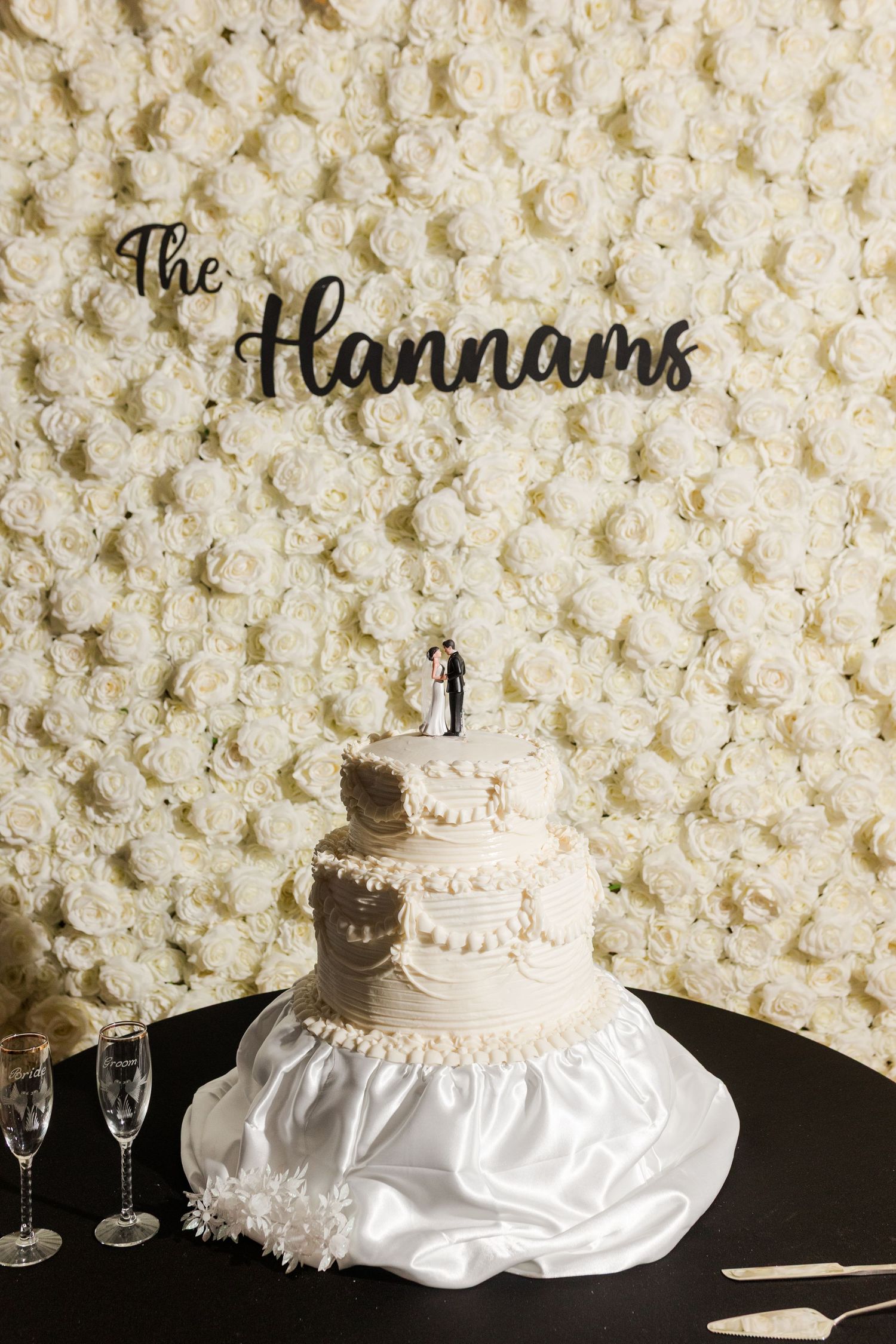 Three-tier white wedding cake with bride and groom topper on a satin-draped table before a cream rose wall with The Hannams sign.