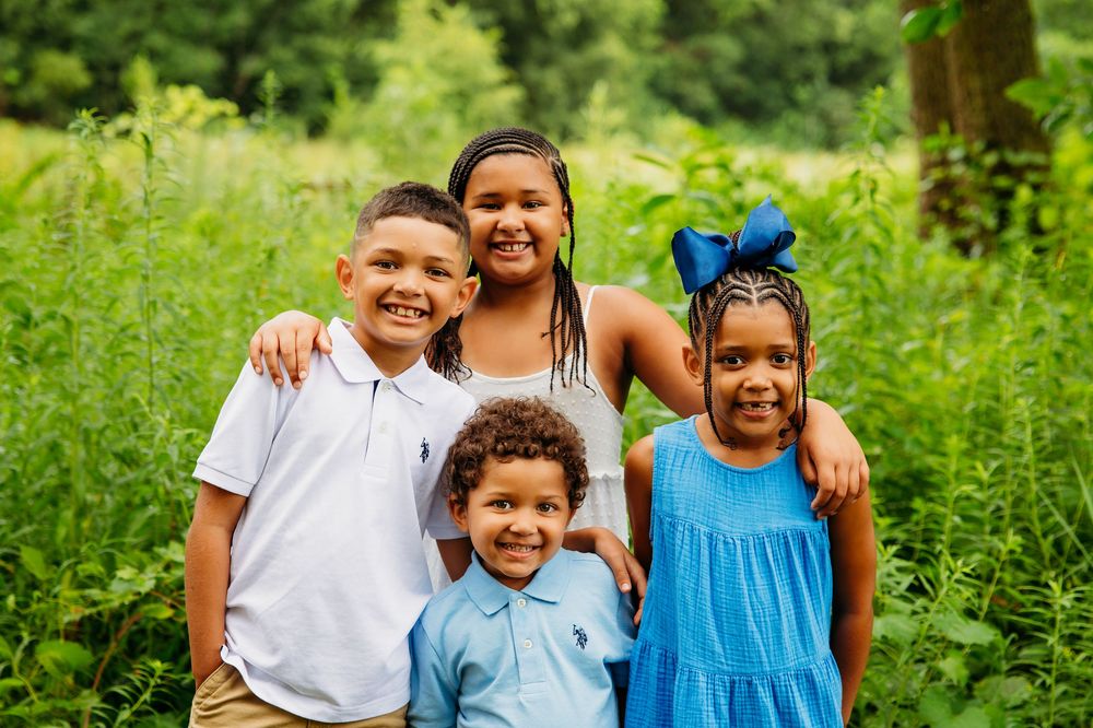 Four siblings smiling together in front of lush green foliage outdoors.