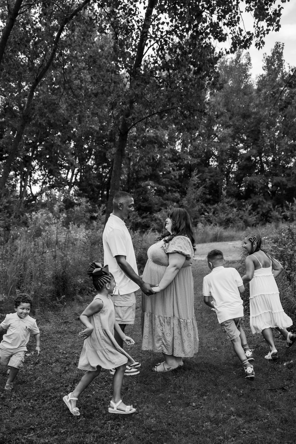 Black and white photo of pregnant couple holding hands surrounded by children.