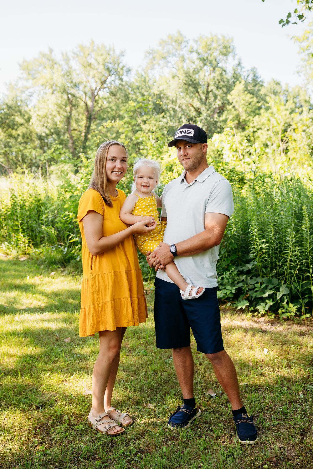 Happy family of three posing together in a lush green outdoor setting.