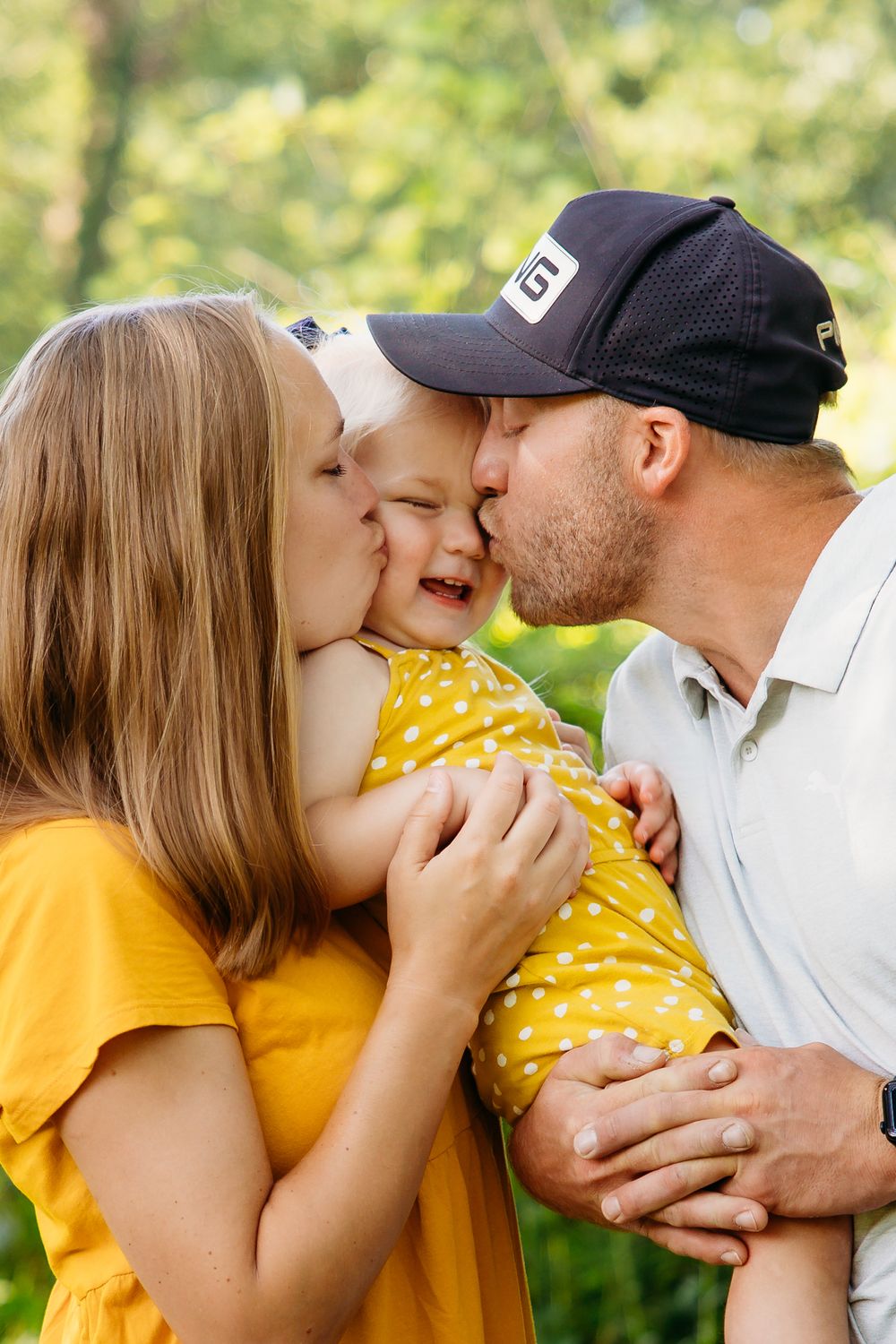 Parents kissing their laughing baby girl on the cheeks outdoors in a park.