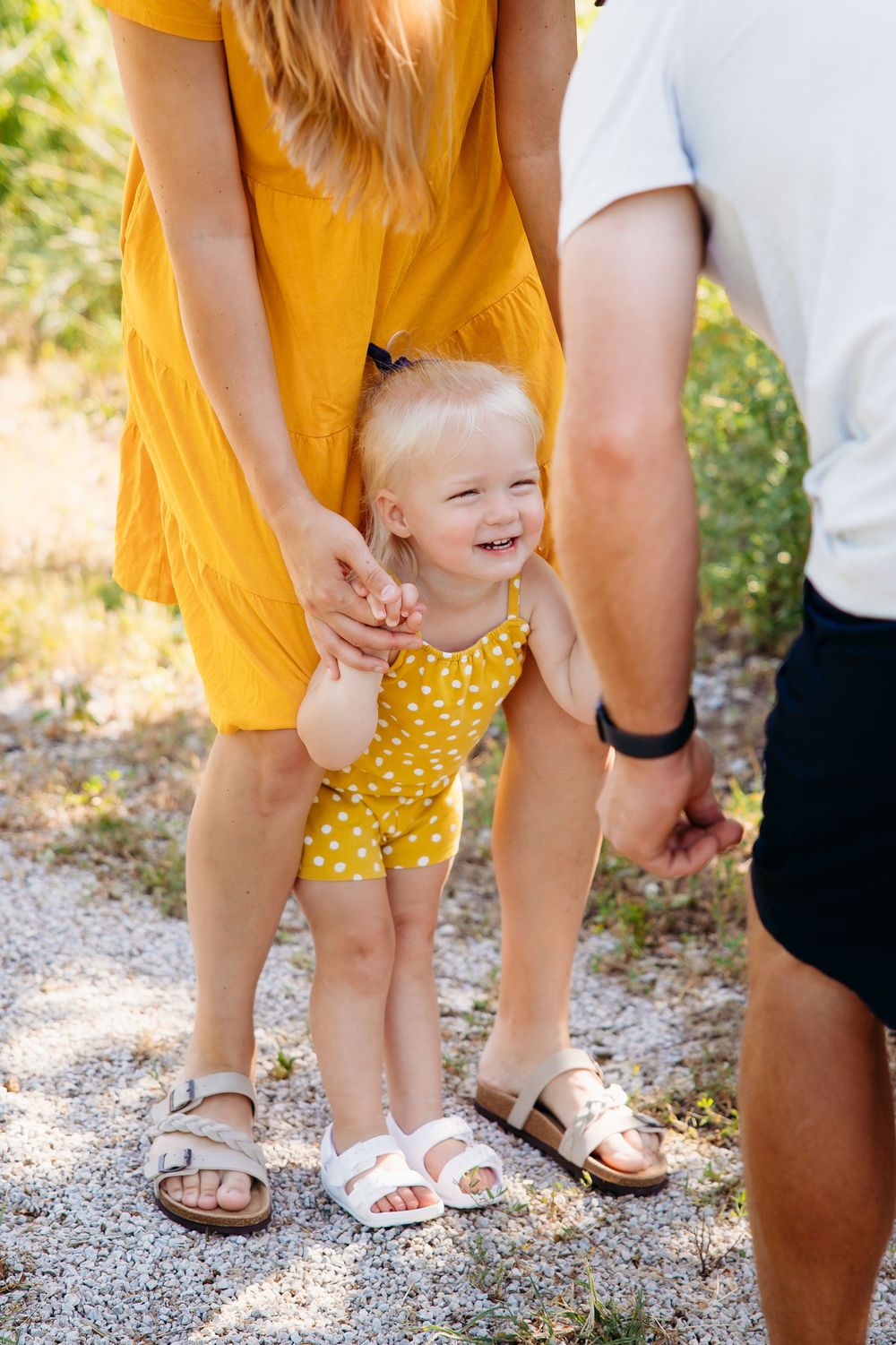 Smiling baby girl in yellow polka dot romper held by parents on a sunny path.