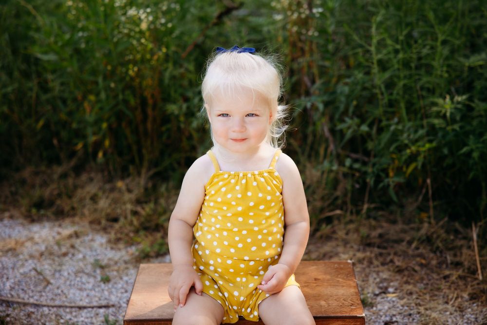 Adorable blonde toddler girl in yellow polka dot romper sitting outdoors.