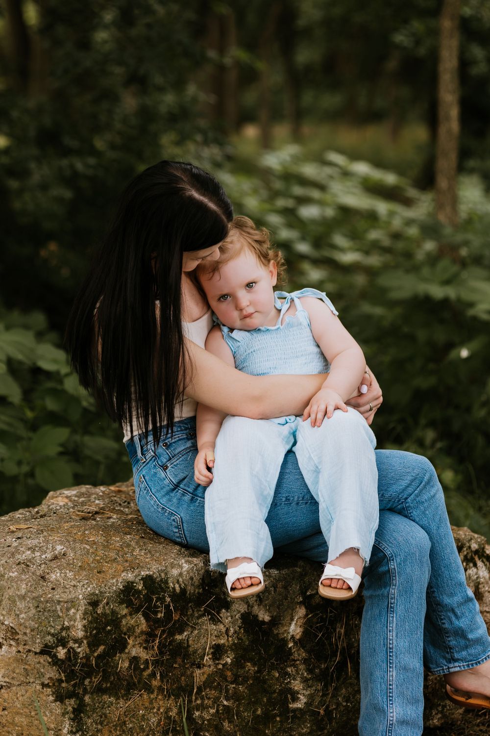 Mother sitting on a stone wall in a forest, tenderly embracing her toddler daughter.