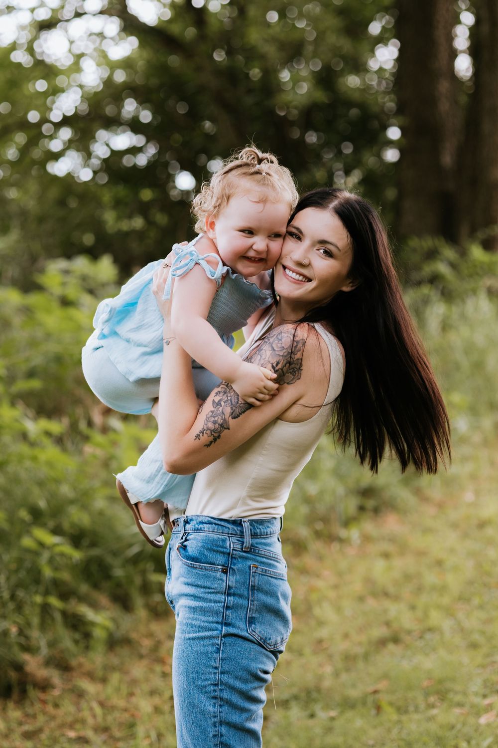 Smiling tattooed mother lifting and hugging her laughing toddler daughter in a green park.
