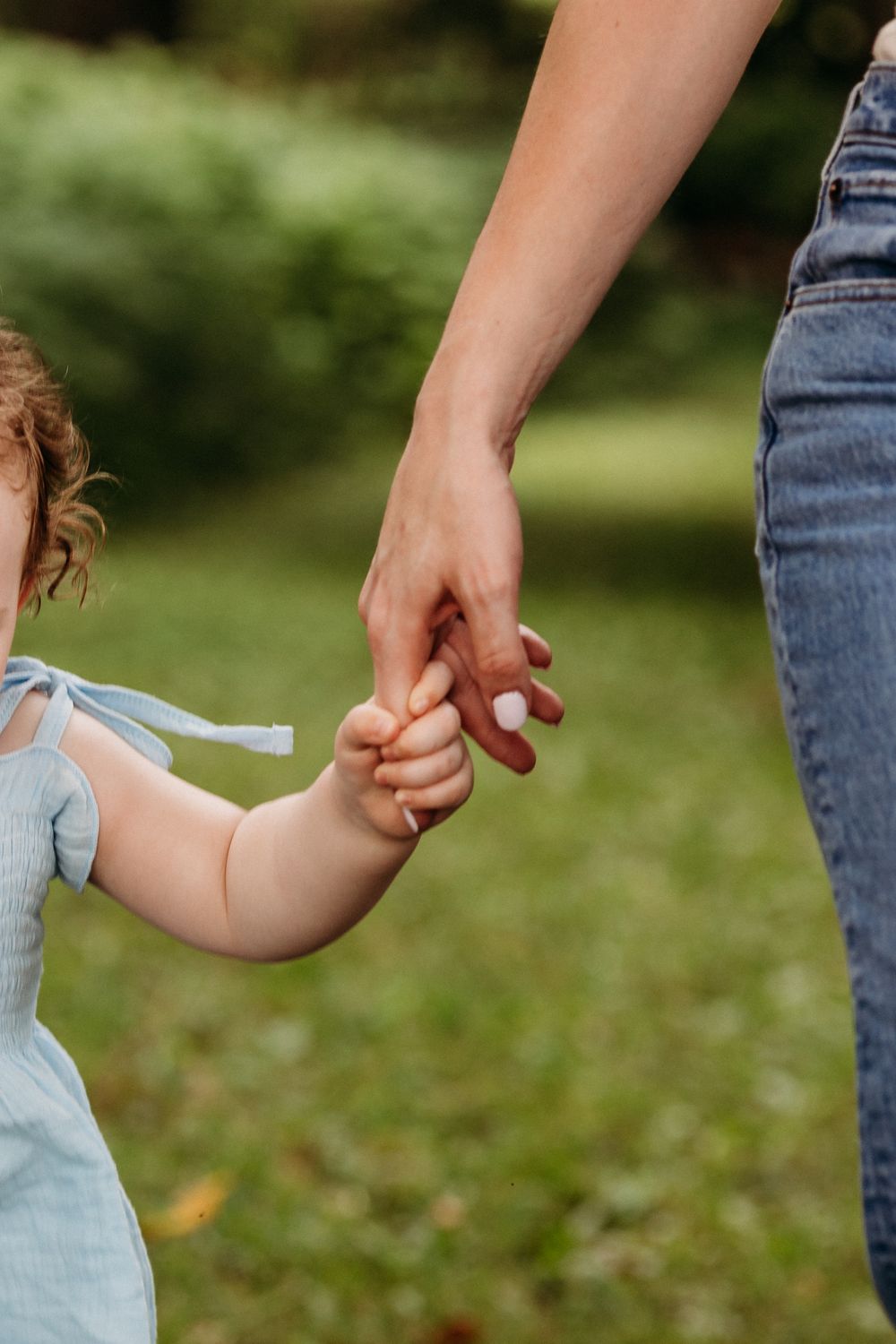 Close-up of a mother and toddler holding hands while walking on green grass outdoors.