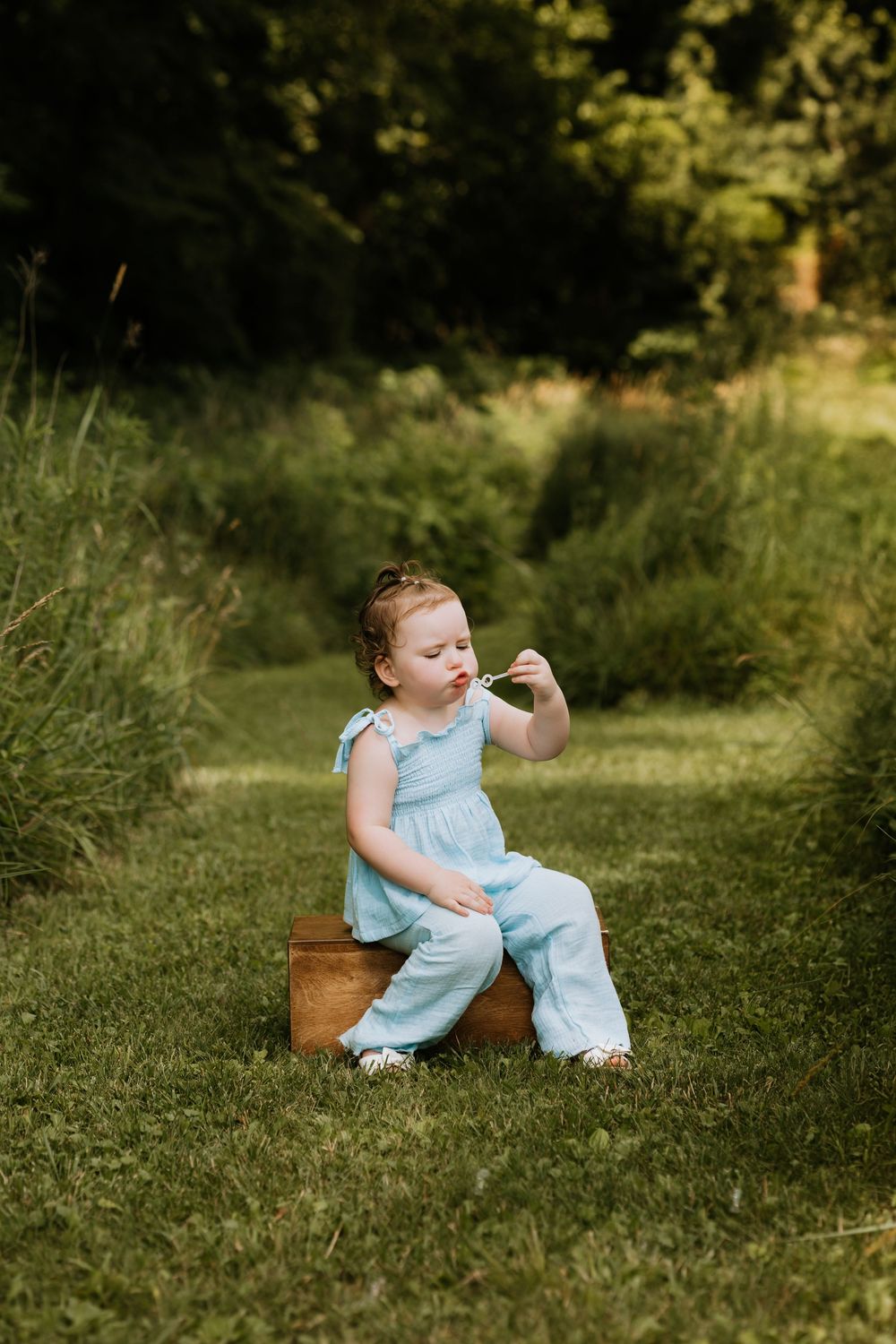 Adorable toddler girl in a light blue jumpsuit sitting on a wooden crate in a garden.