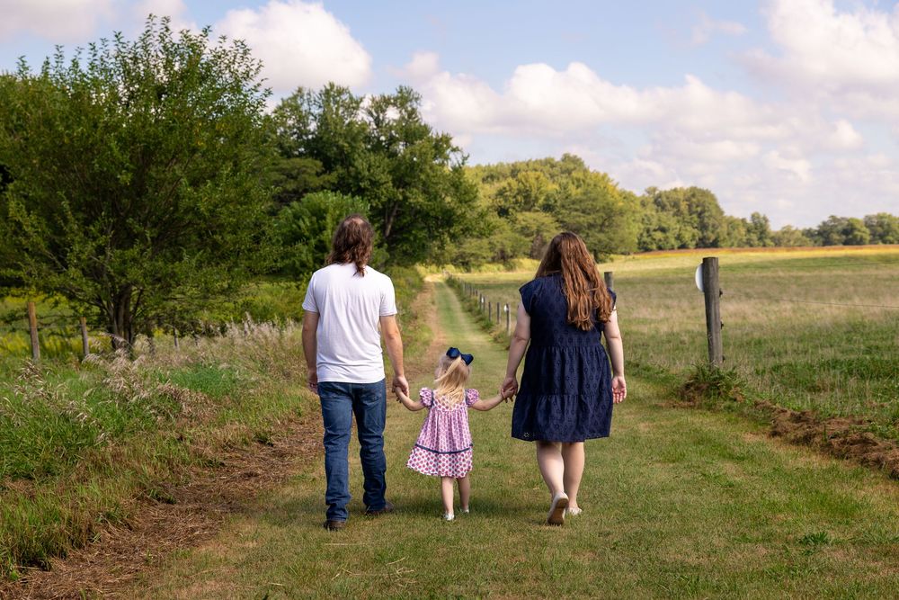 Family of three walking hand in hand down a scenic green path in the countryside.
