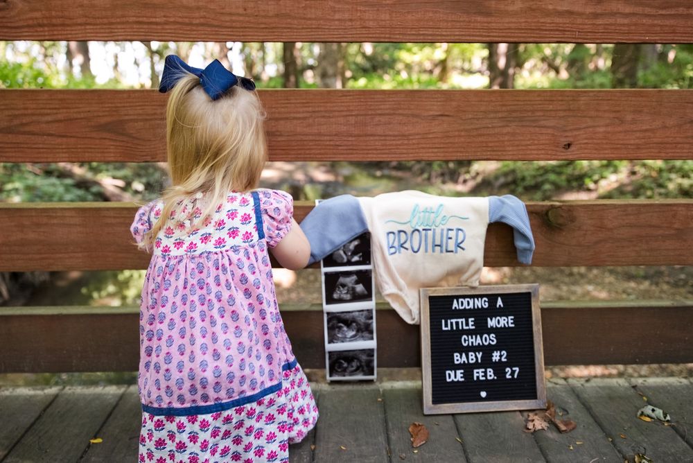 Toddler girl with pregnancy announcement sign and baby brother onesie on a bench.