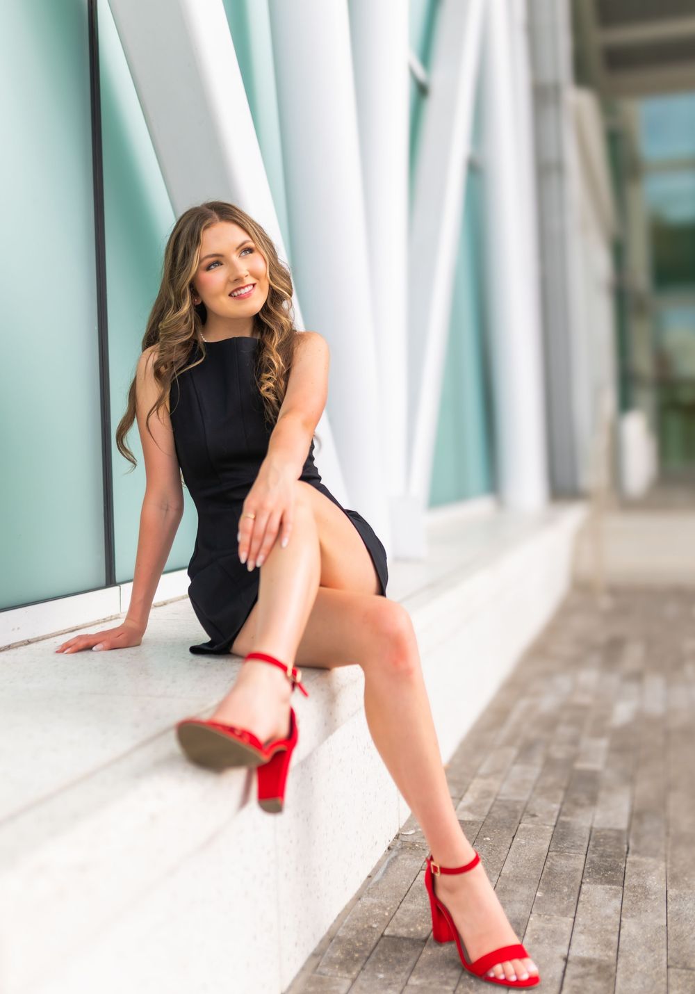 Young woman in black mini dress and red heels sitting on steps outdoors smiling.