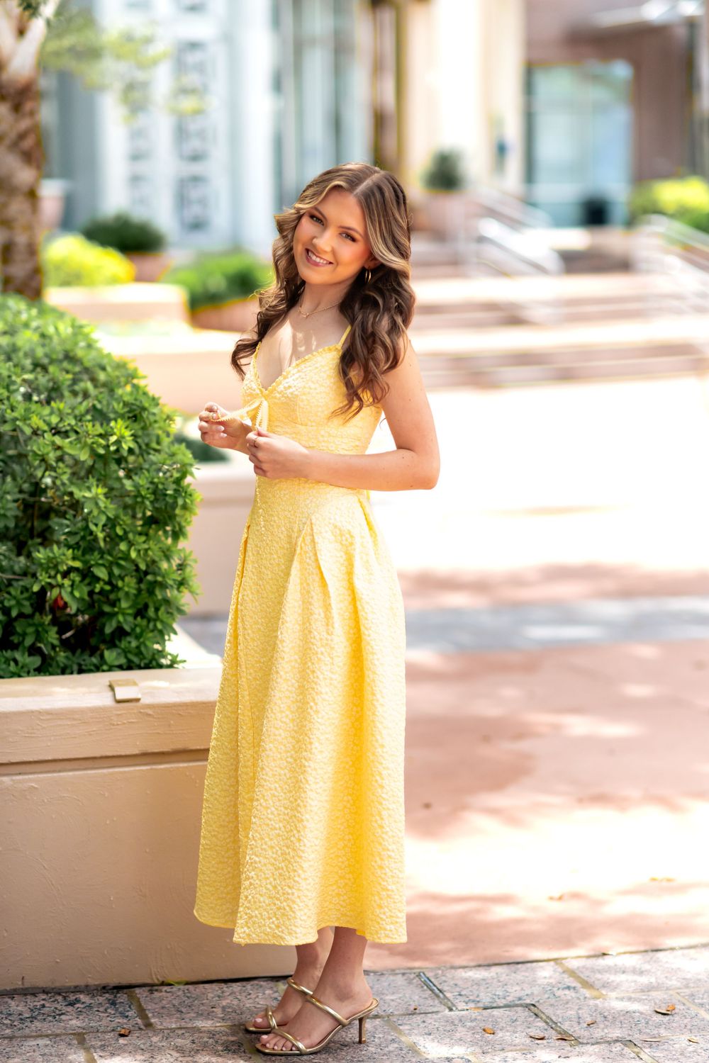 Young woman smiling in a yellow midi dress, posing outdoors near greenery in a sunny urban setting.