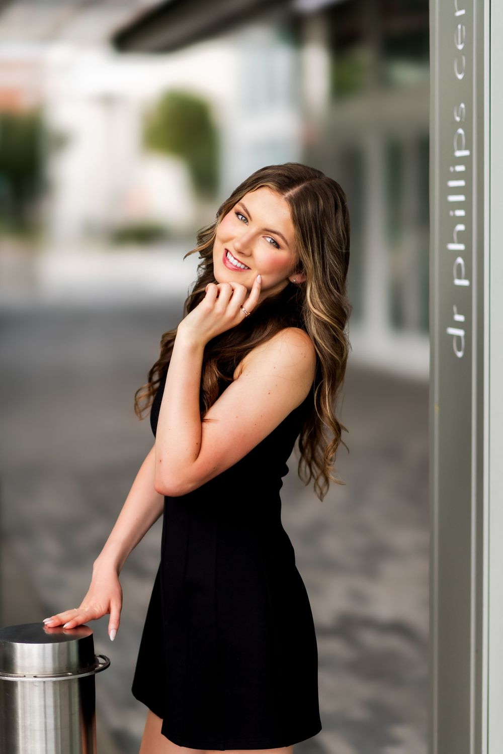 Young woman in black dress smiling outdoors near Dr. Phillips Center, posing gracefully with hand near face.