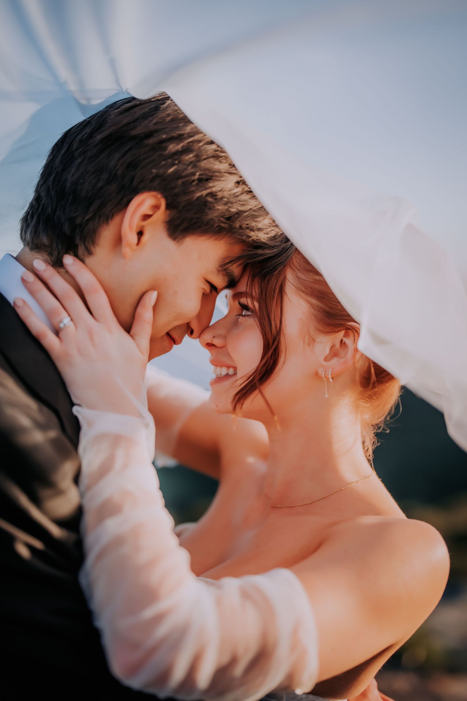 A bride and groom share a romantic moment under a flowing white veil, smiling at each other on their wedding day.