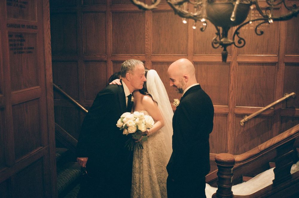 Father of the bride kisses daughter in wedding dress at elegant wood-paneled venue while groom stands nearby.