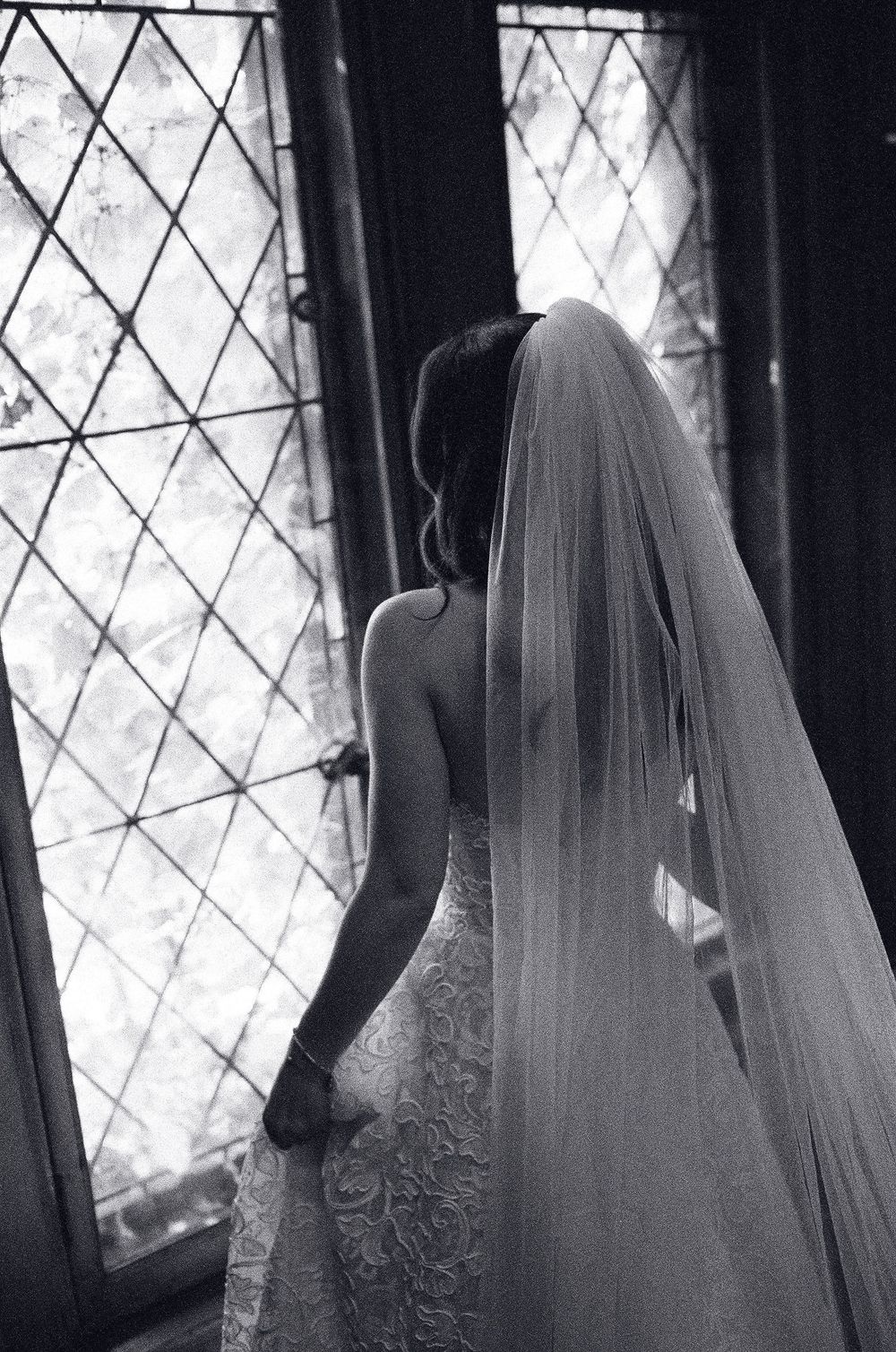 Black and white photo of a bride in lace gown and long veil standing before diamond-paned windows.