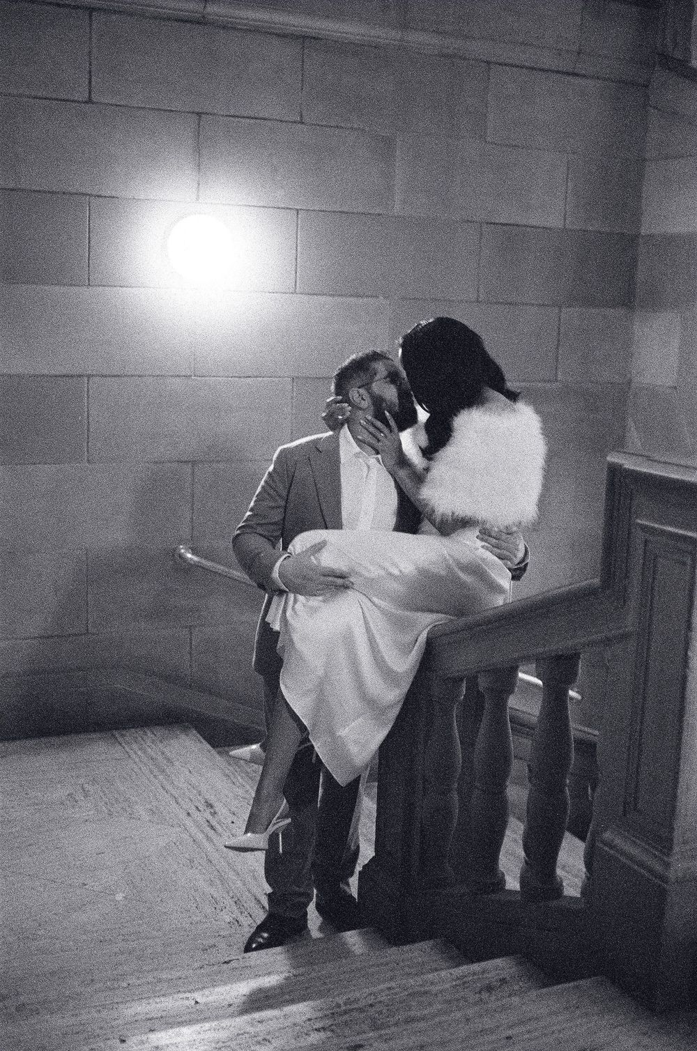Black and white photo of a couple sharing a romantic kiss on a staircase railing, with a bright light glowing behind them.