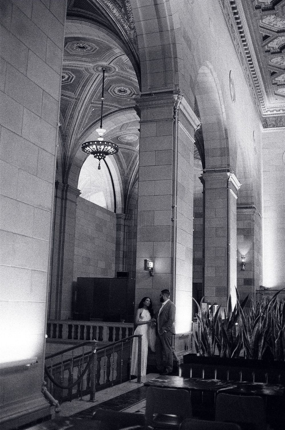 Couple stands beneath ornate arched ceilings and chandeliers in a grand historic building interior, black and white photo.