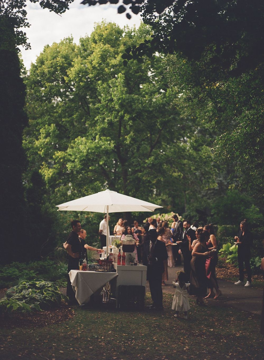 Guests mingle at an outdoor garden party with a white umbrella bar setup surrounded by lush green trees.