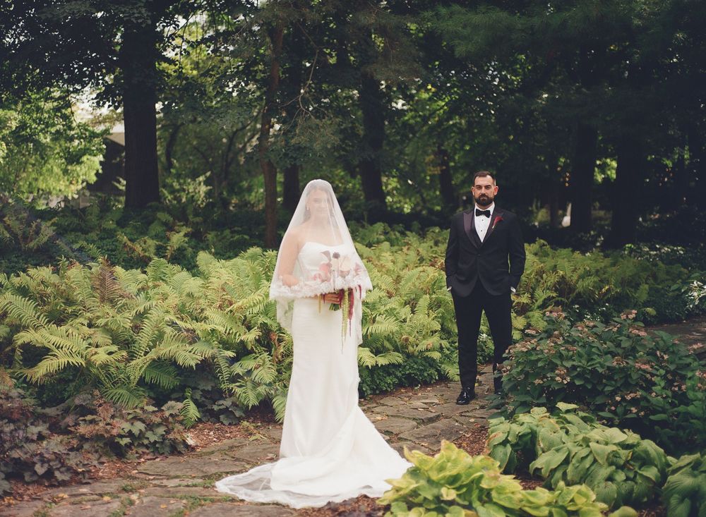 Bride in white gown and veil stands with groom in black tuxedo in a lush green garden wedding setting.