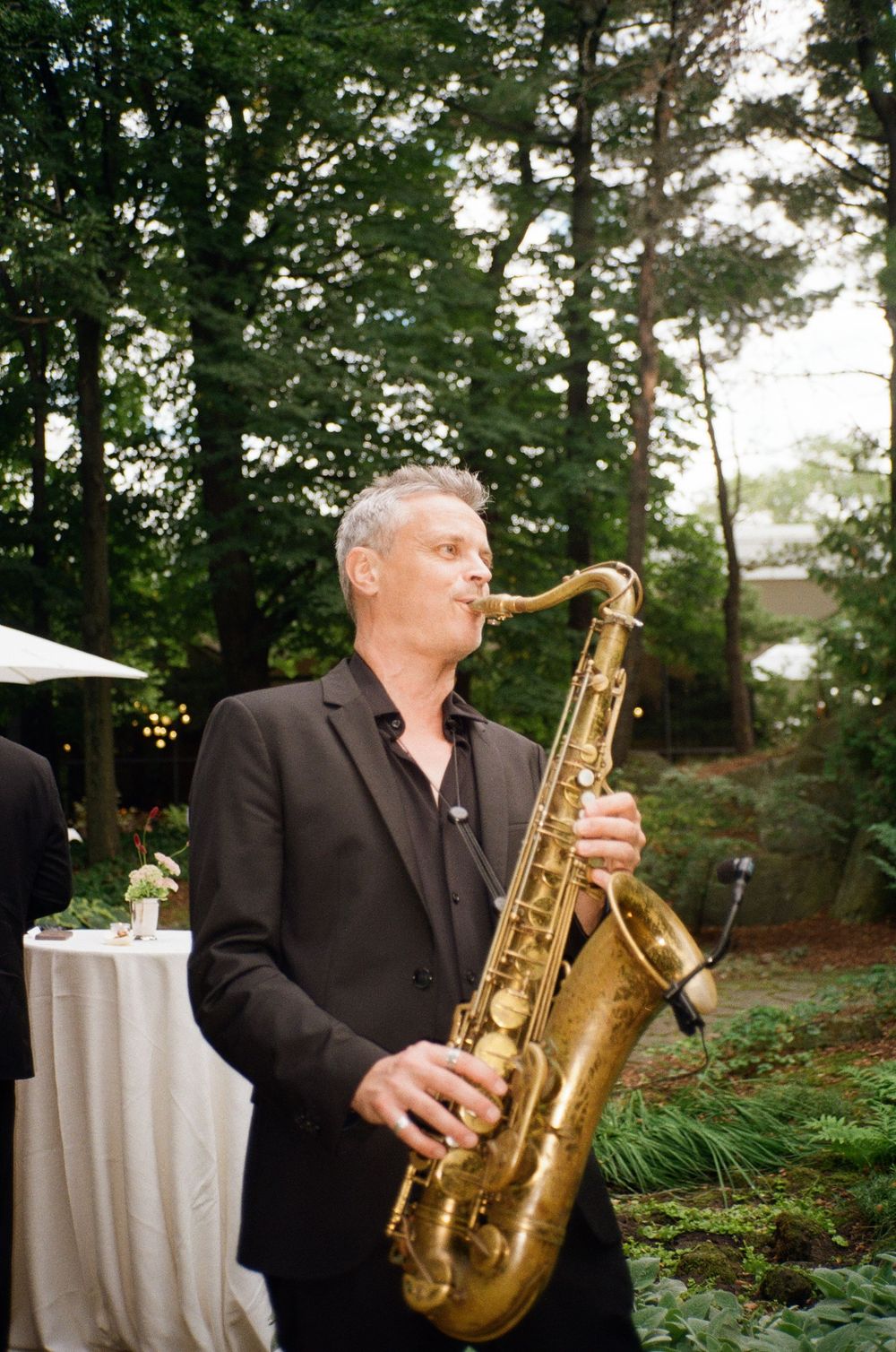 A man in a black suit plays a gold saxophone outdoors at an elegant garden event surrounded by tall trees.
