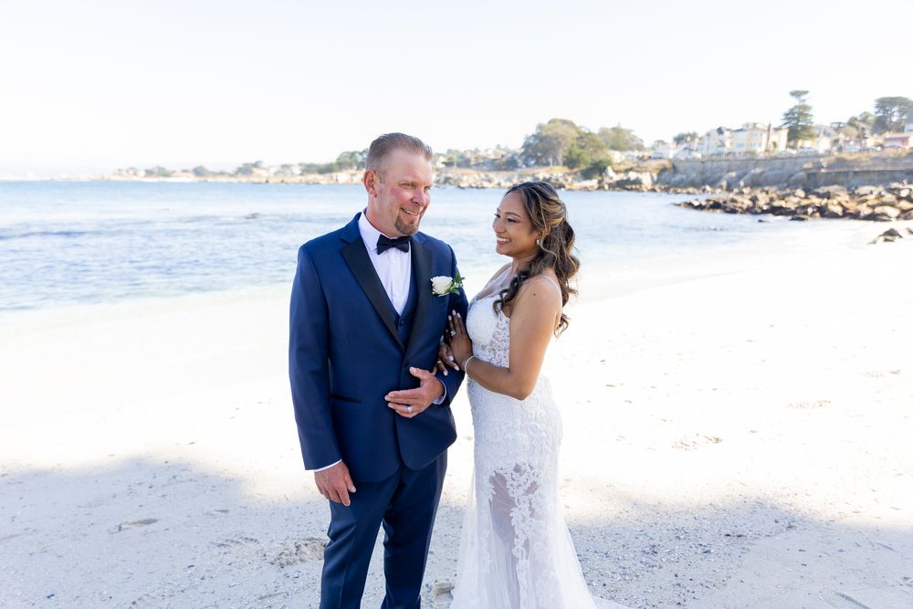 Bride and groom smiling at each other on a sunny beach, groom in navy suit, bride in white lace gown.