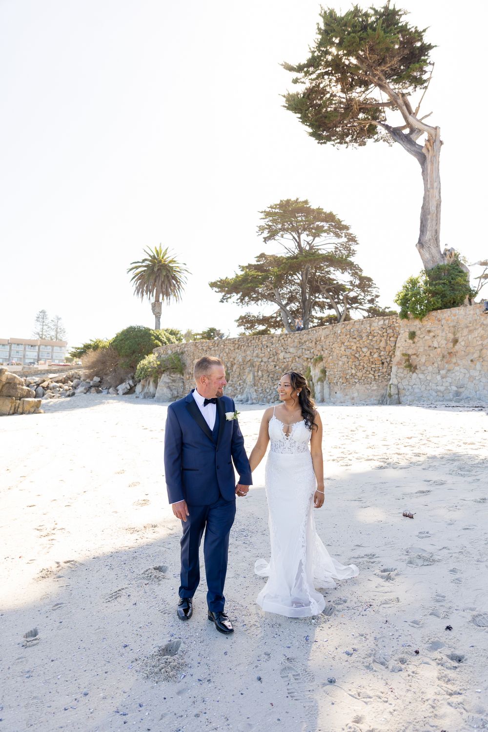 Bride and groom hold hands walking on sandy beach, groom in navy suit, bride in white gown, trees and stone wall behind.