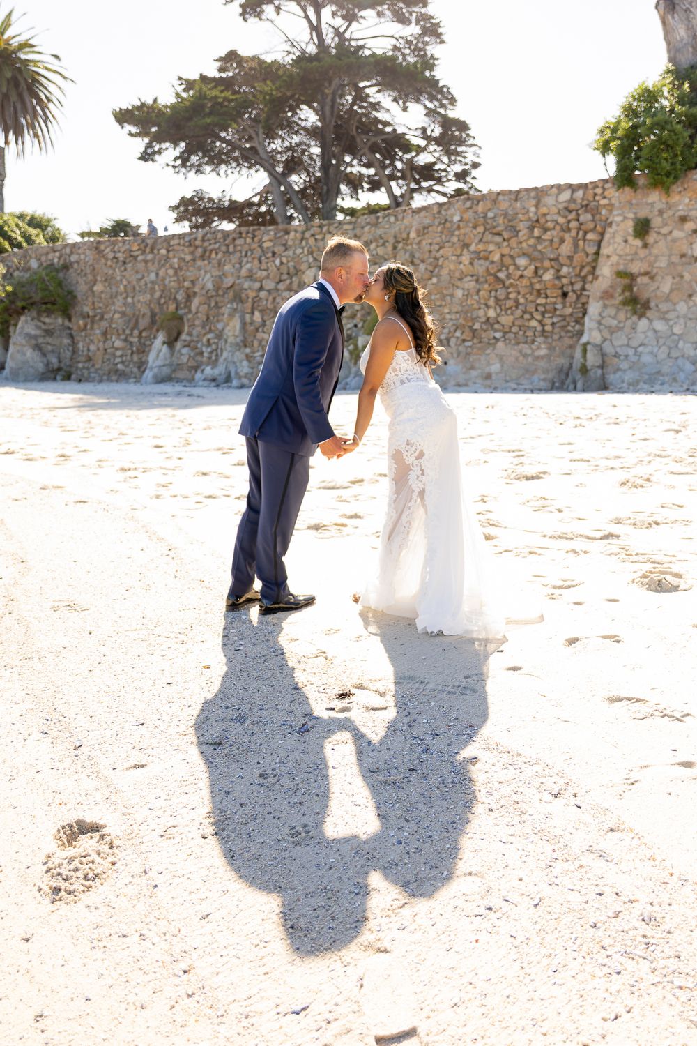 Bride and groom kissing on sandy beach, casting shadows, stone wall and trees in background on sunny wedding day.