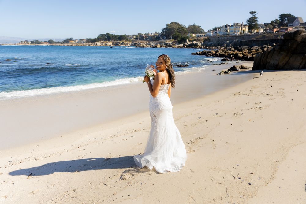 Bride in white lace wedding gown standing on sandy beach with ocean waves and coastal town in background.