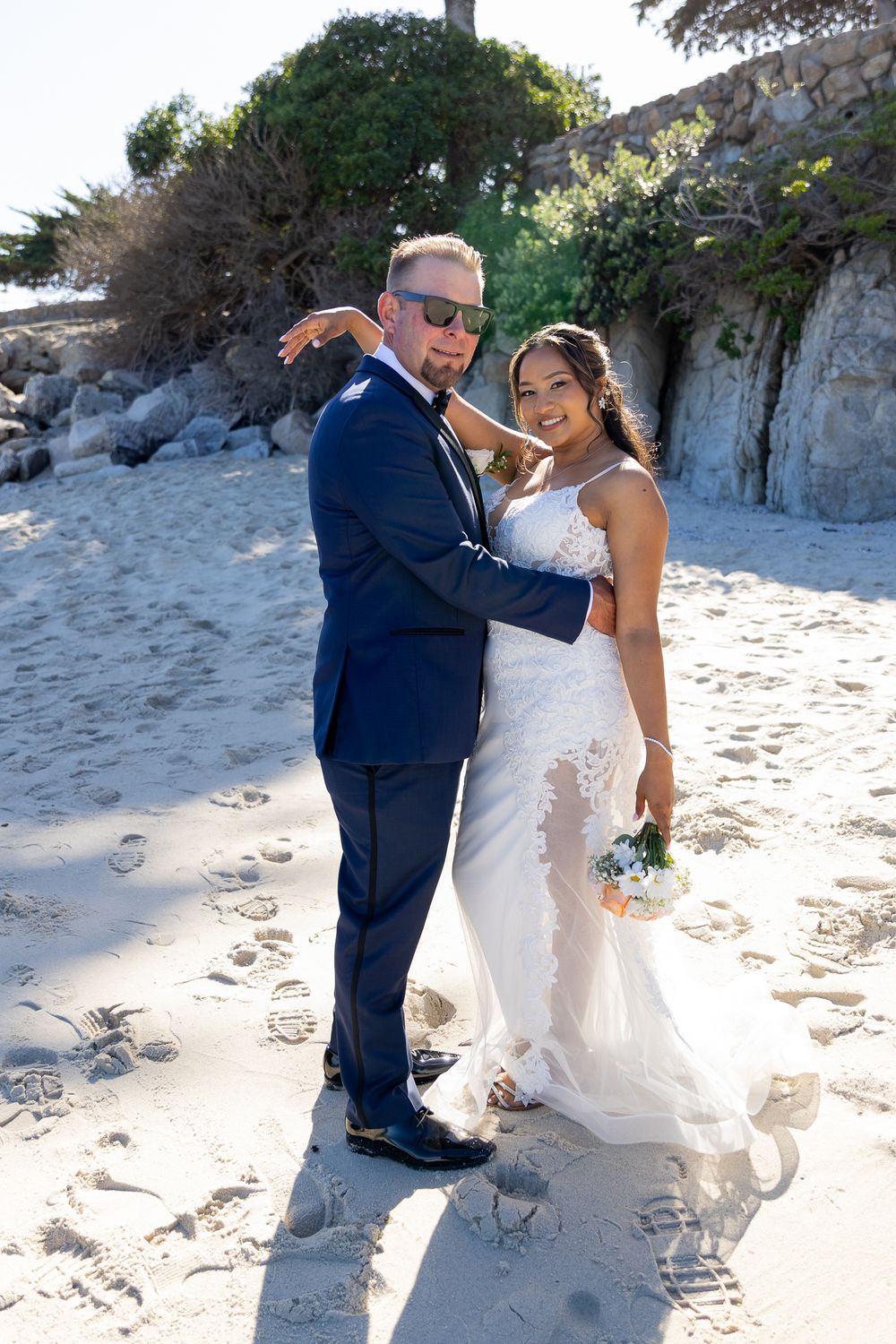 Bride and groom posing on sandy beach, she holds white bouquet in lace gown, he wears navy suit with sunglasses.