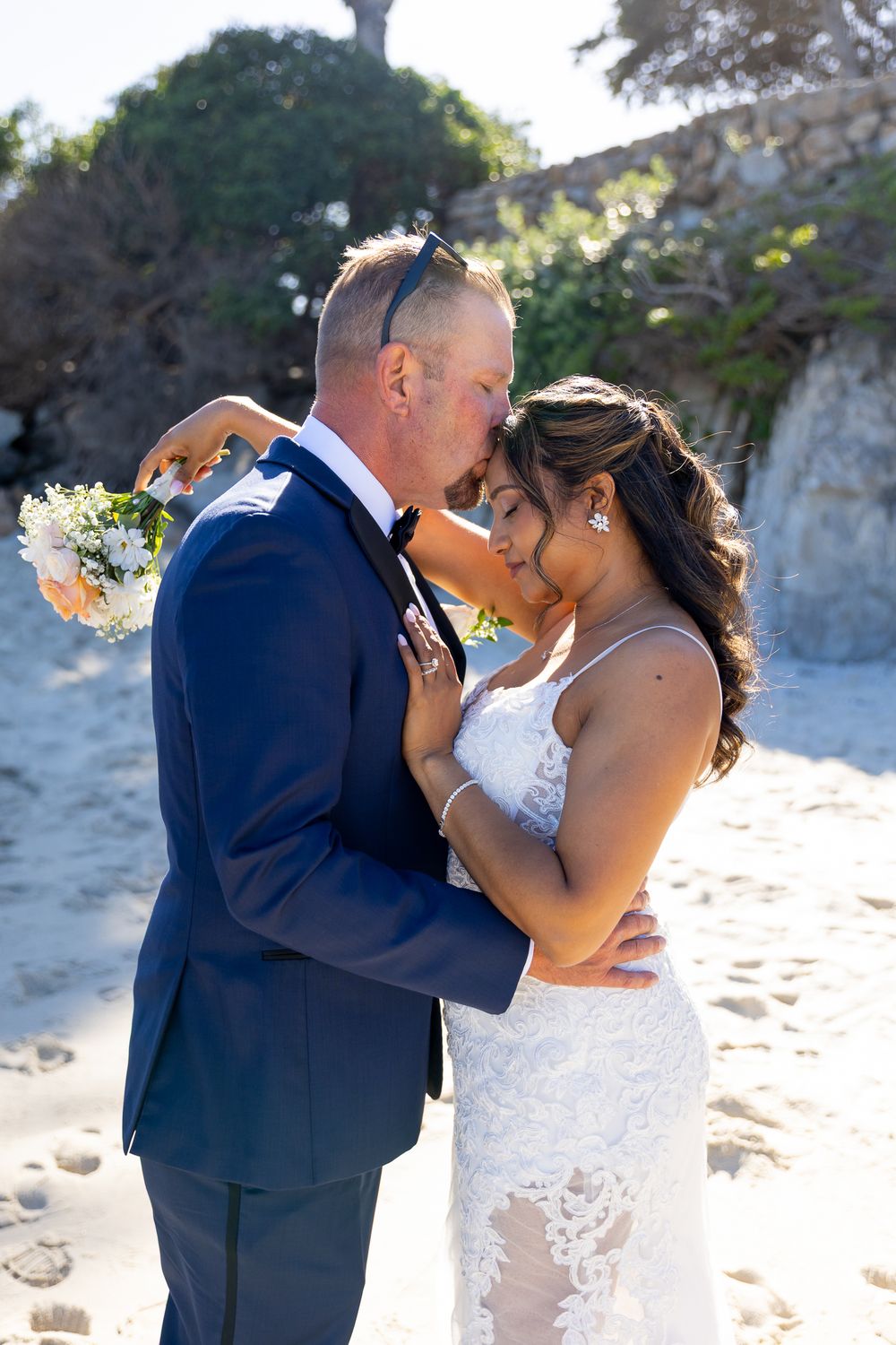 Couple sharing romantic kiss on beach, groom in navy suit holding bouquet, bride in lace wedding dress.