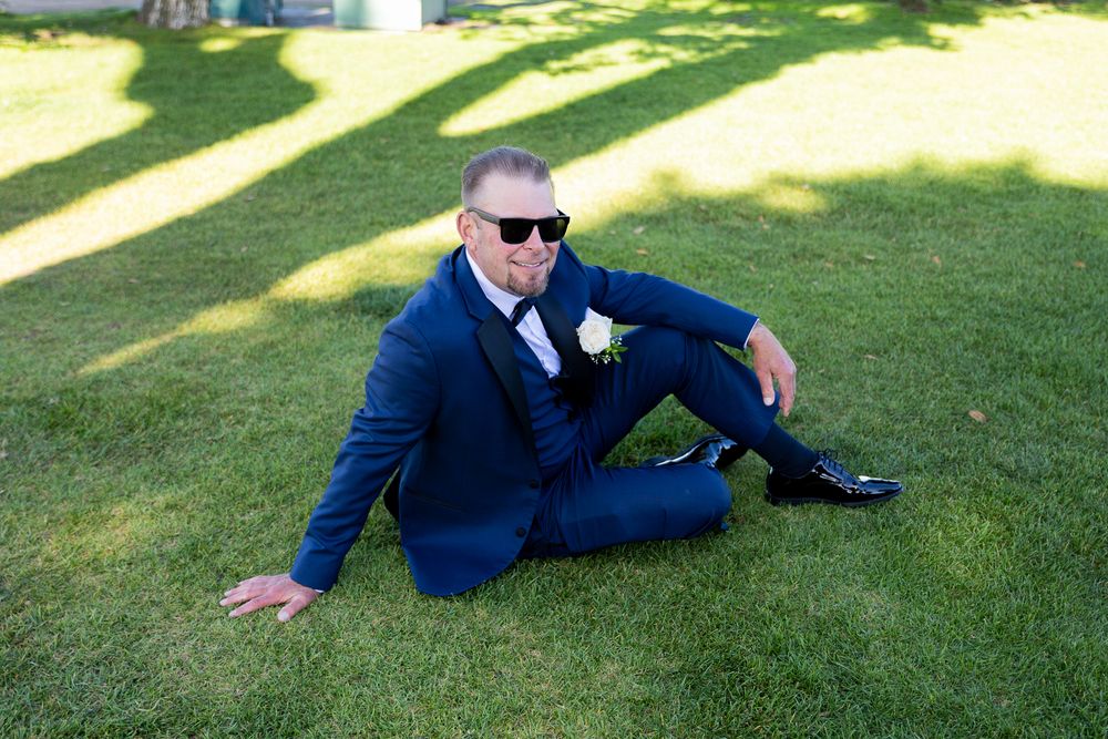 Man in blue suit and sunglasses sitting on green grass with white boutonniere, posing for wedding photo.