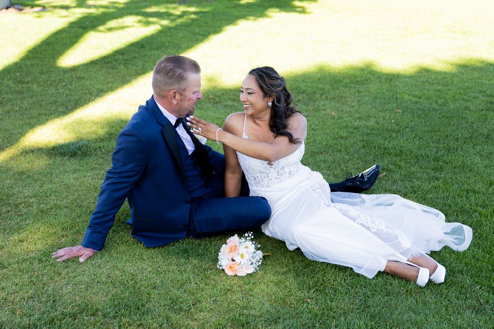 Bride and groom sitting on green grass, smiling at each other, with a bouquet nearby on their wedding day.
