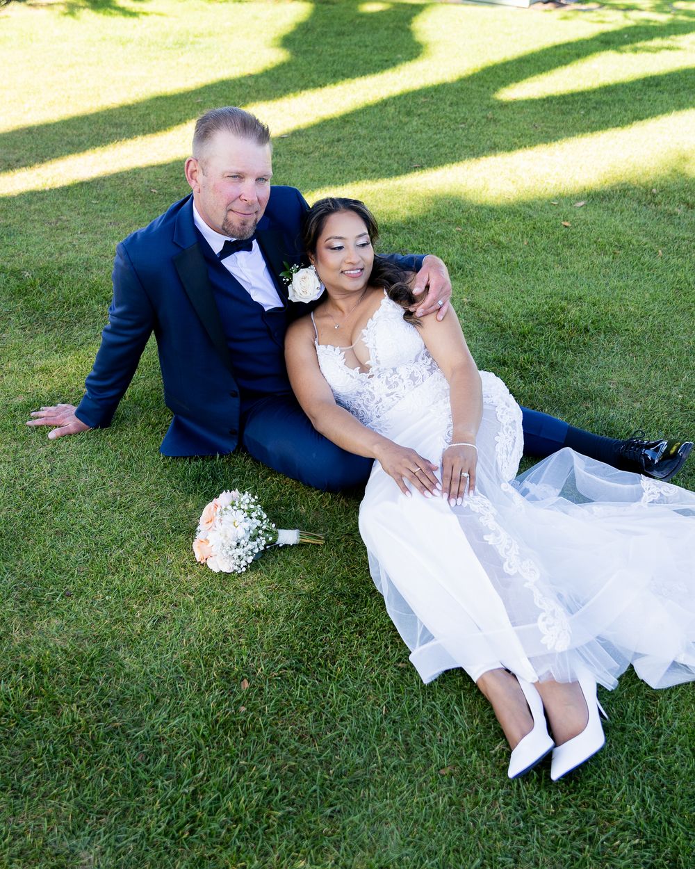 Bride and groom sitting on green grass, bride in white lace gown holding bouquet, groom in navy suit with bow tie.