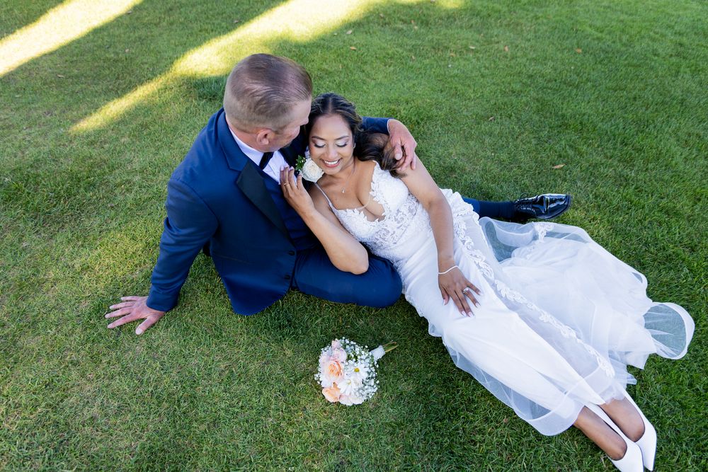 Bride and groom sitting on green grass, sharing a romantic moment in wedding attire, with a bridal bouquet nearby.