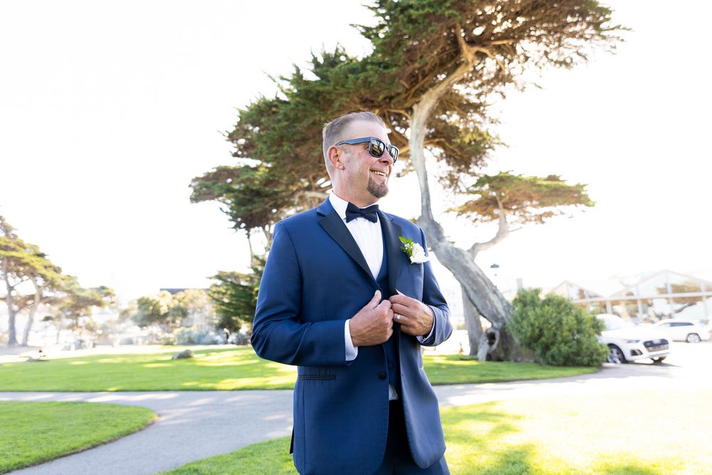 Man in blue tuxedo with bow tie and boutonniere wearing sunglasses outdoors at a wedding ceremony.