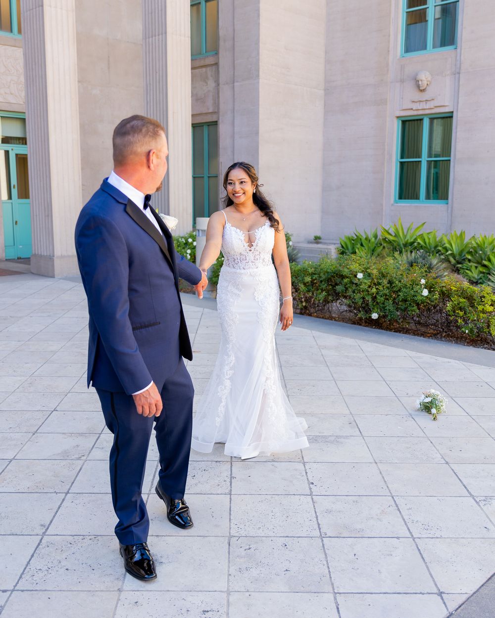Bride in white mermaid gown shares first look moment with groom in navy suit outside elegant building.