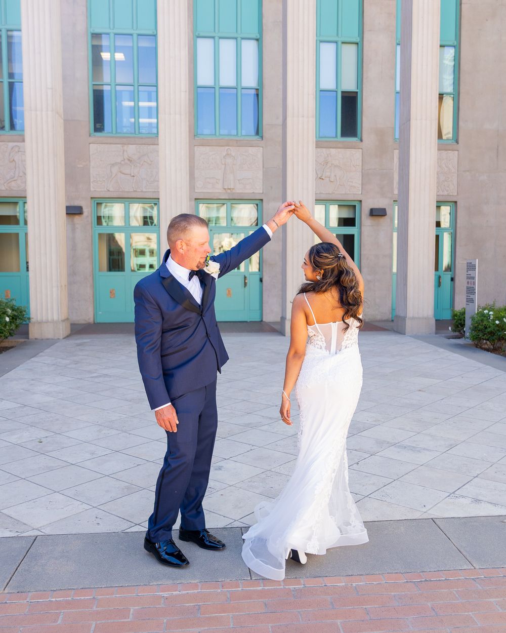 Bride and groom dancing outdoors in front of a building, man in navy suit twirling woman in white wedding gown.