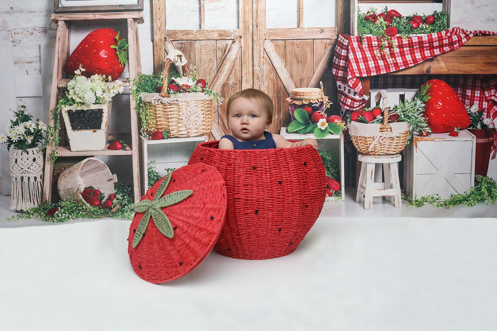 Cute baby sitting inside a giant red strawberry prop in a colorful farm-themed photo studio.