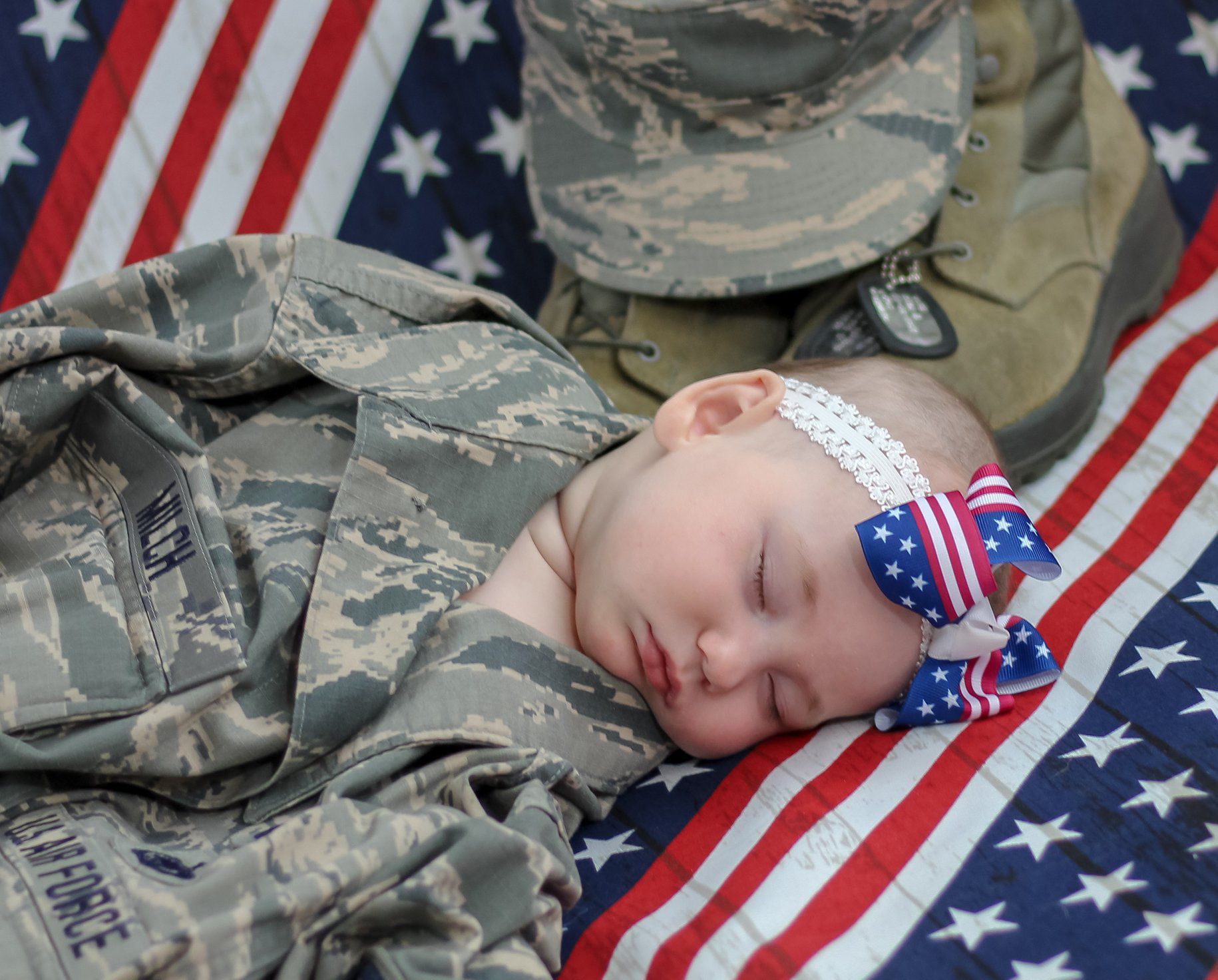 Sleeping newborn baby wrapped in military camouflage with American flag bow headband on US flag backdrop.