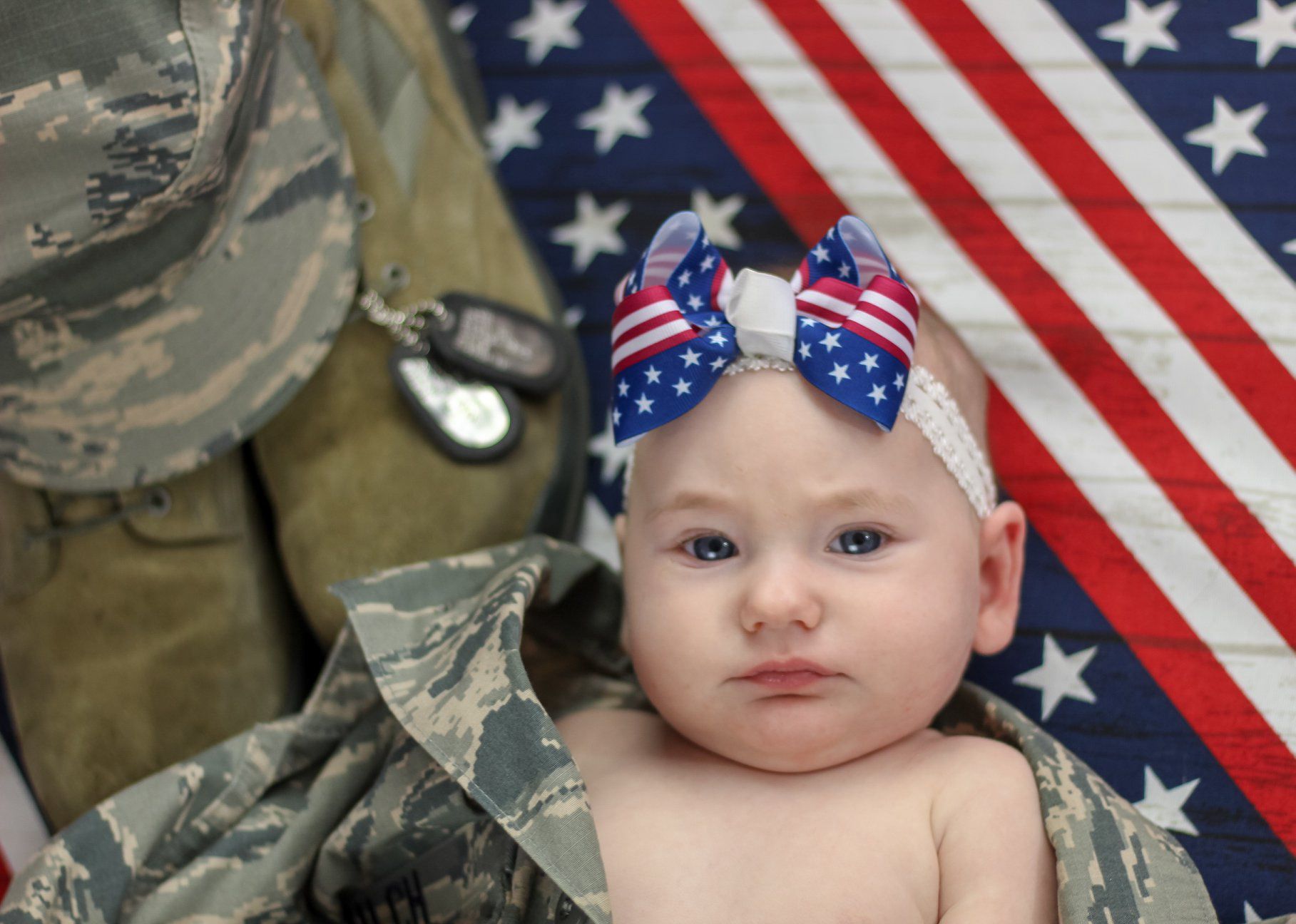 Newborn baby with patriotic American flag bow headband lying on military camouflage uniform with dog tags.
