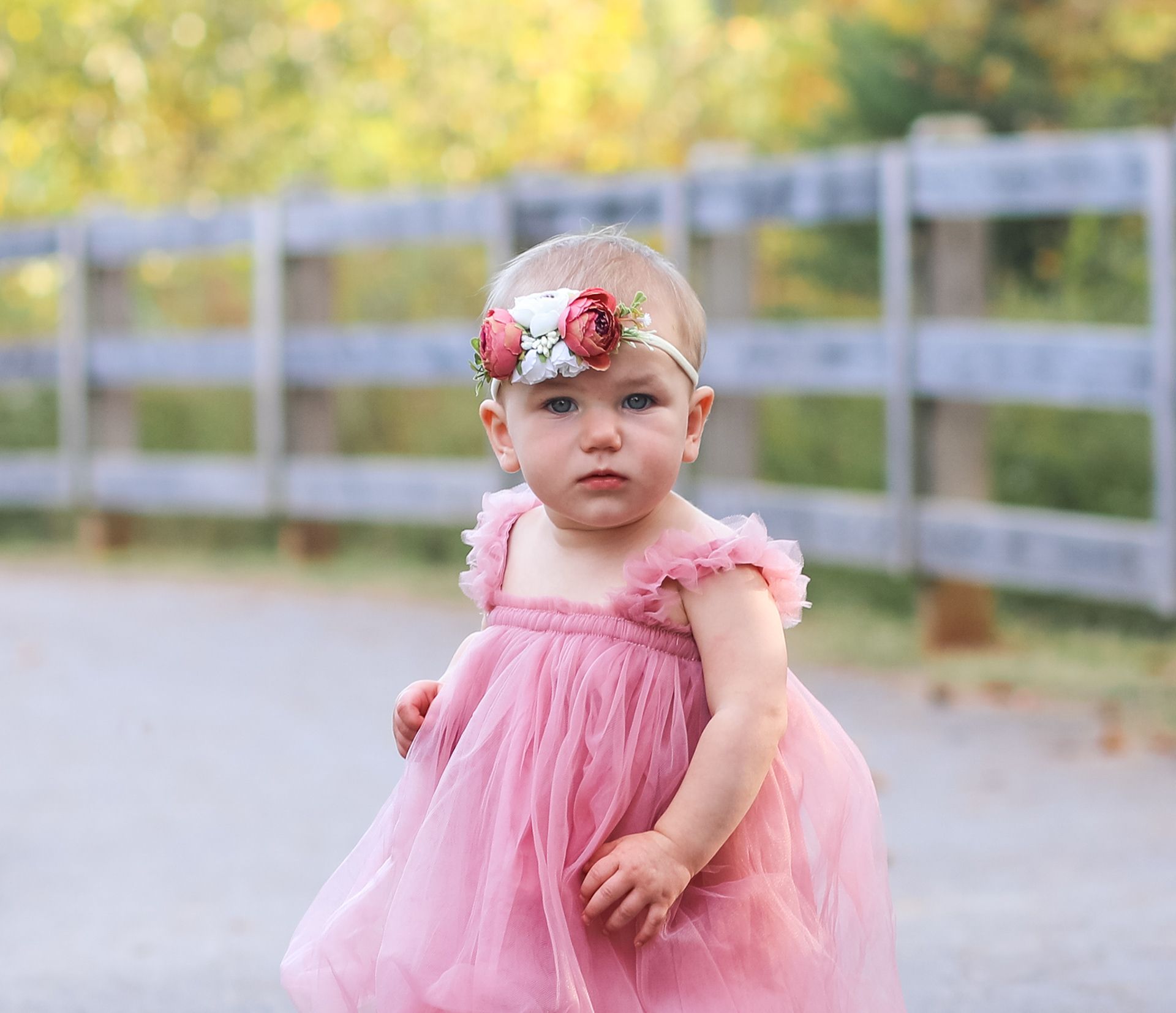 A baby girl in a pink tulle dress and floral headband poses outdoors near a wooden fence.