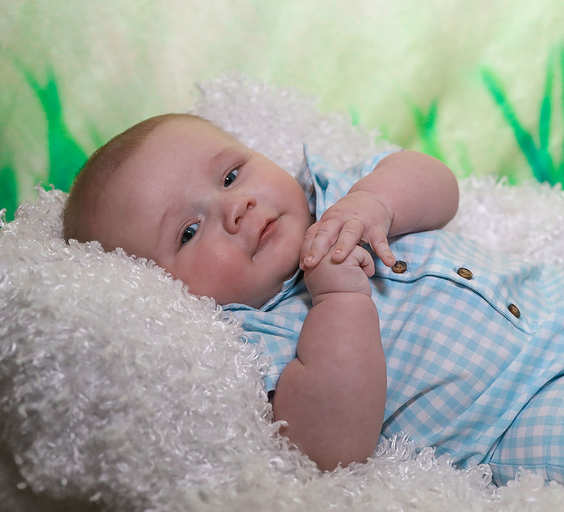 Adorable infant in light blue checkered outfit resting on white textured blanket looking at camera.