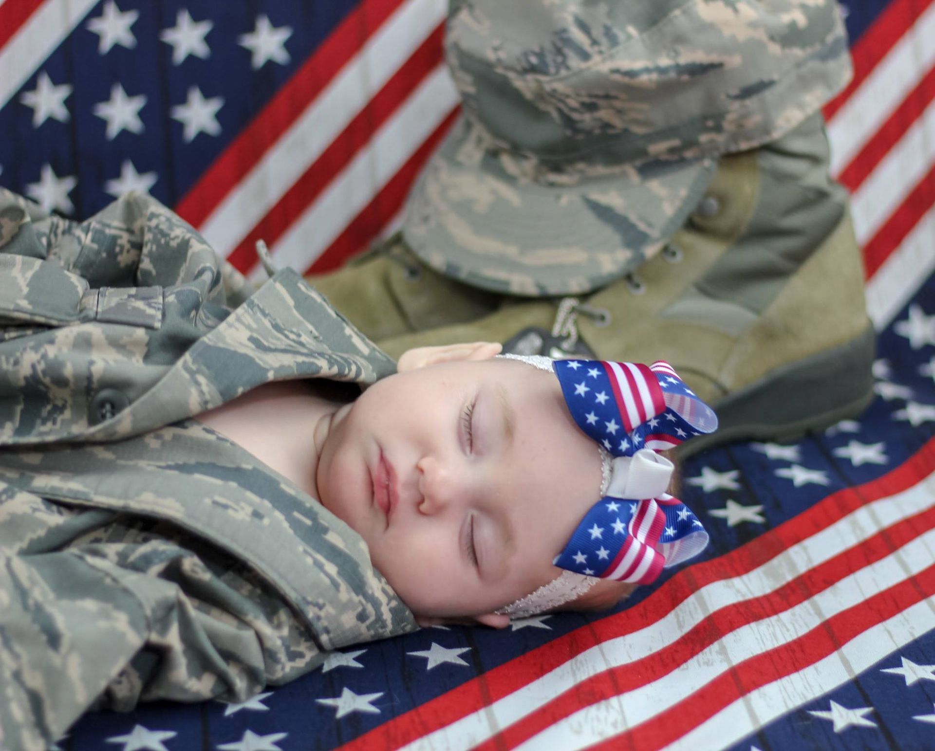 Newborn baby sleeping on American flag with patriotic bow headband beside military boots and uniform.