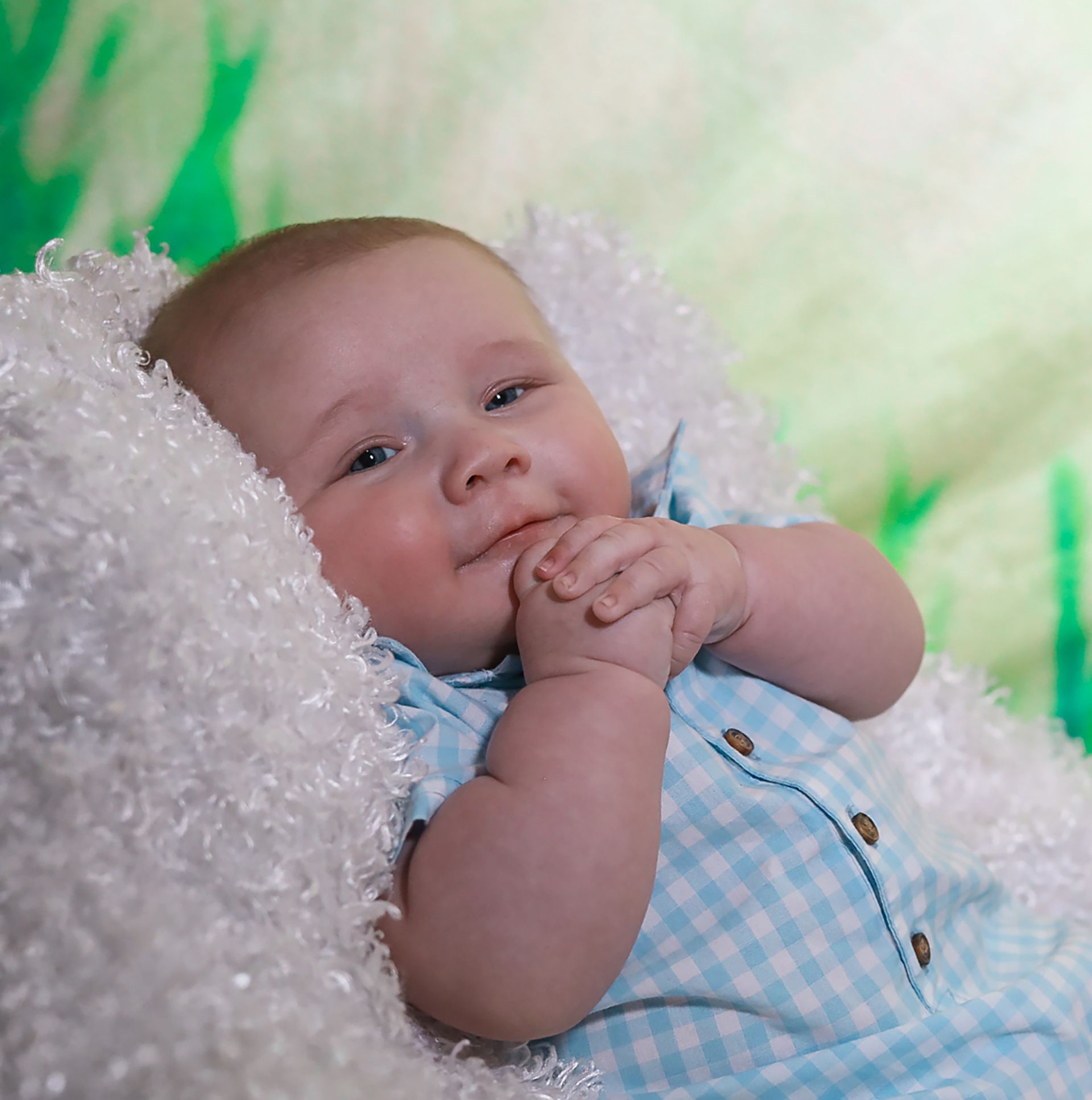 Sweet baby boy in blue gingham romper lying on white fluffy blanket with green background.