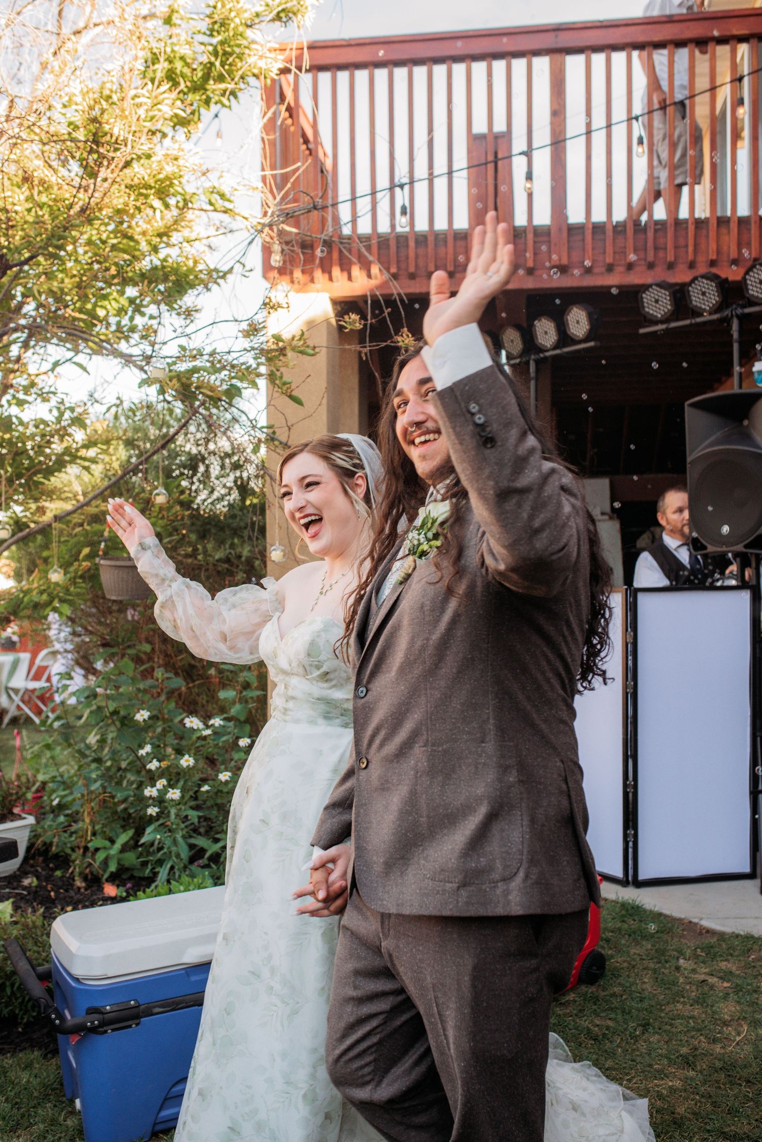 Joyful newlyweds celebrate their backyard wedding reception, raising hands in excitement, bride in lace gown and groom in gray suit.