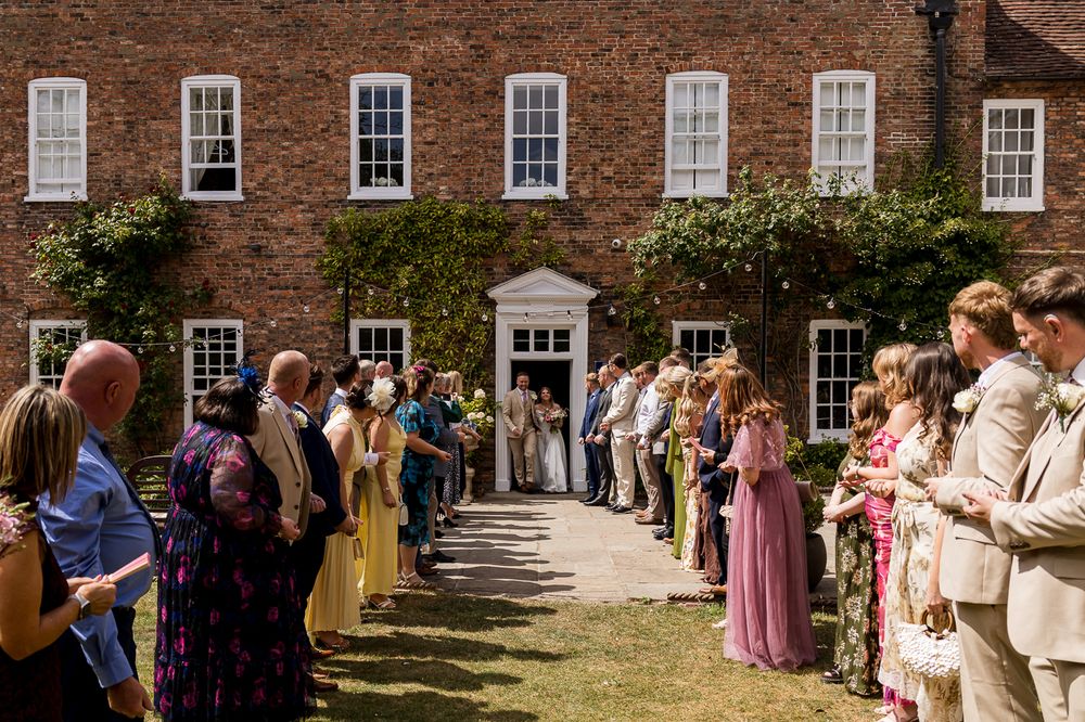 Wedding guests line a sunny garden path outside a Georgian brick manor, awaiting the bride and groom.